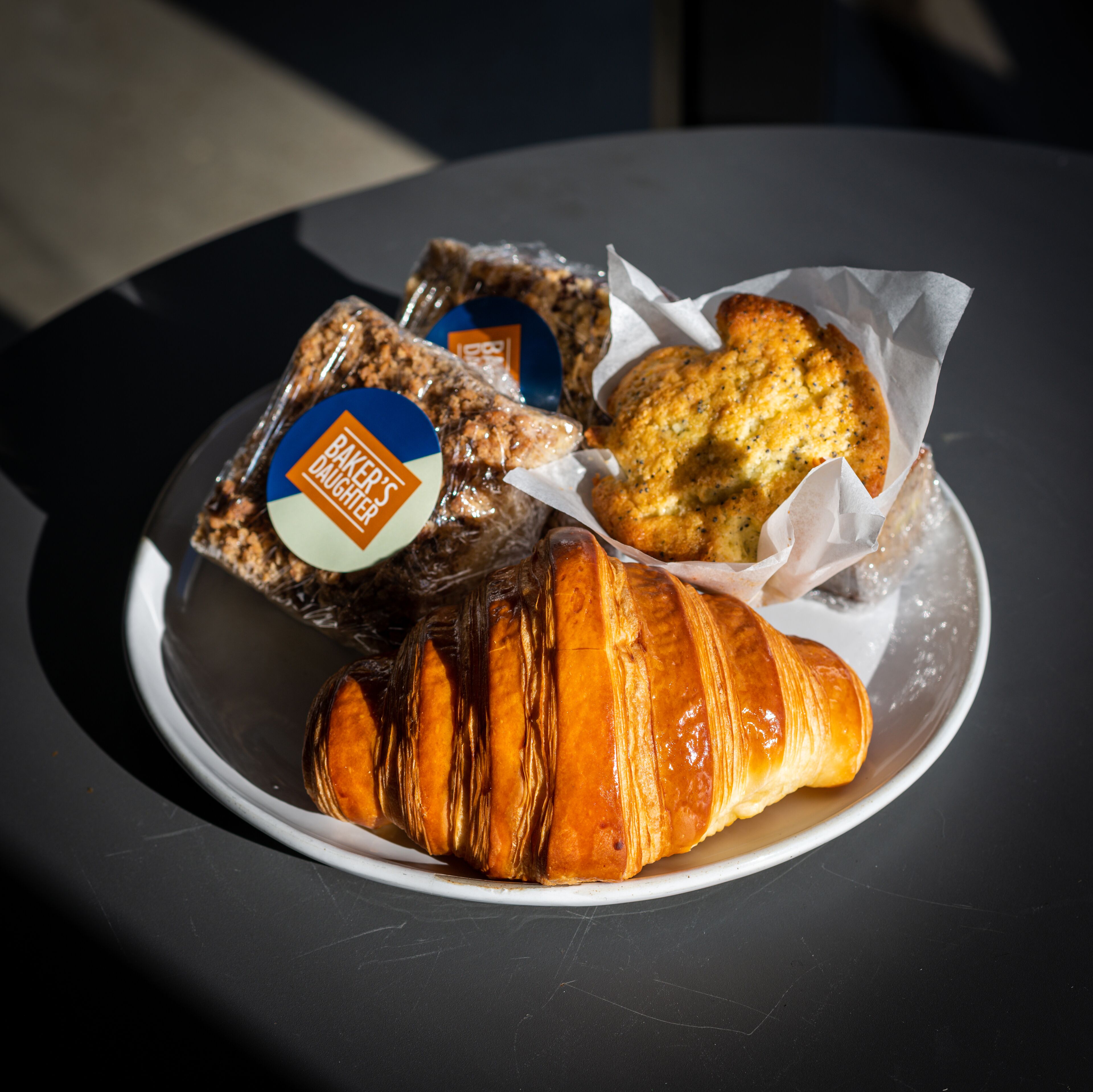 Ein Teller mit einem Croissant, einem Muffin und einer verpackten Zwischenmahlzeit auf einem Tisch.