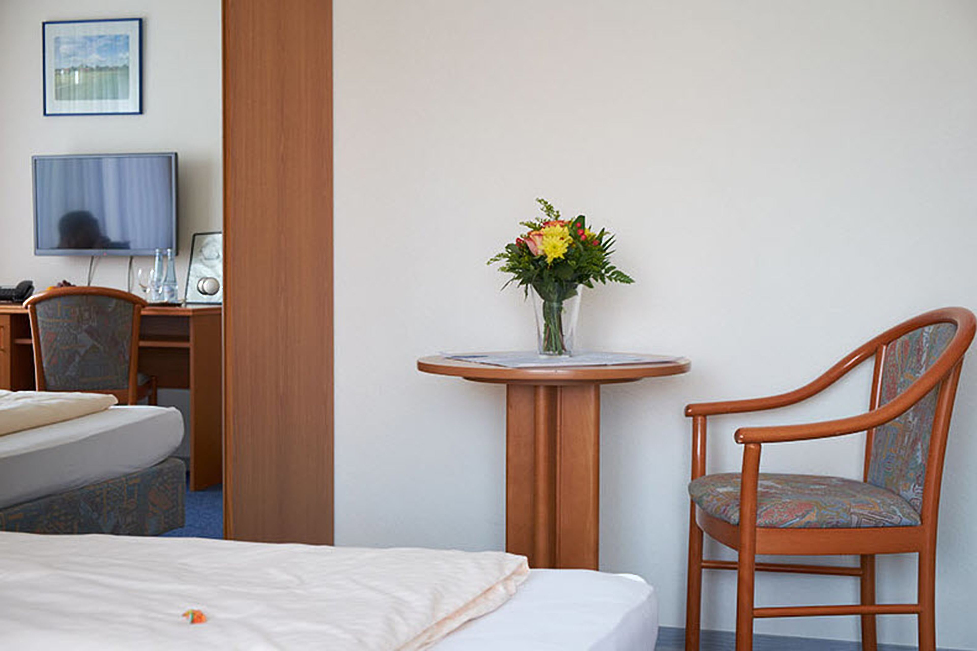 A hotel room with a bed, a desk with a computer, a chair, and a small table with a vase of flowers.