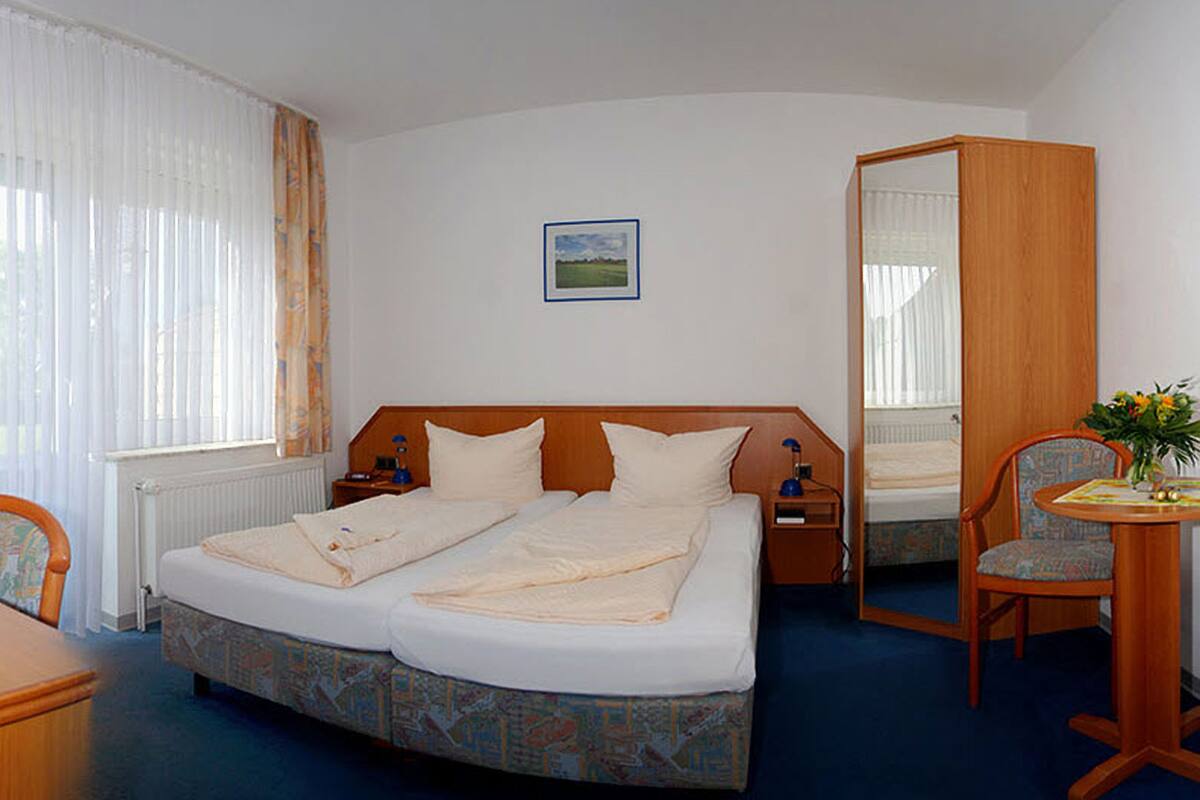 A hotel room with a double bed, a wooden headboard, a bedside table with a lamp, a chair, a small table with a vase, and a window with curtains.