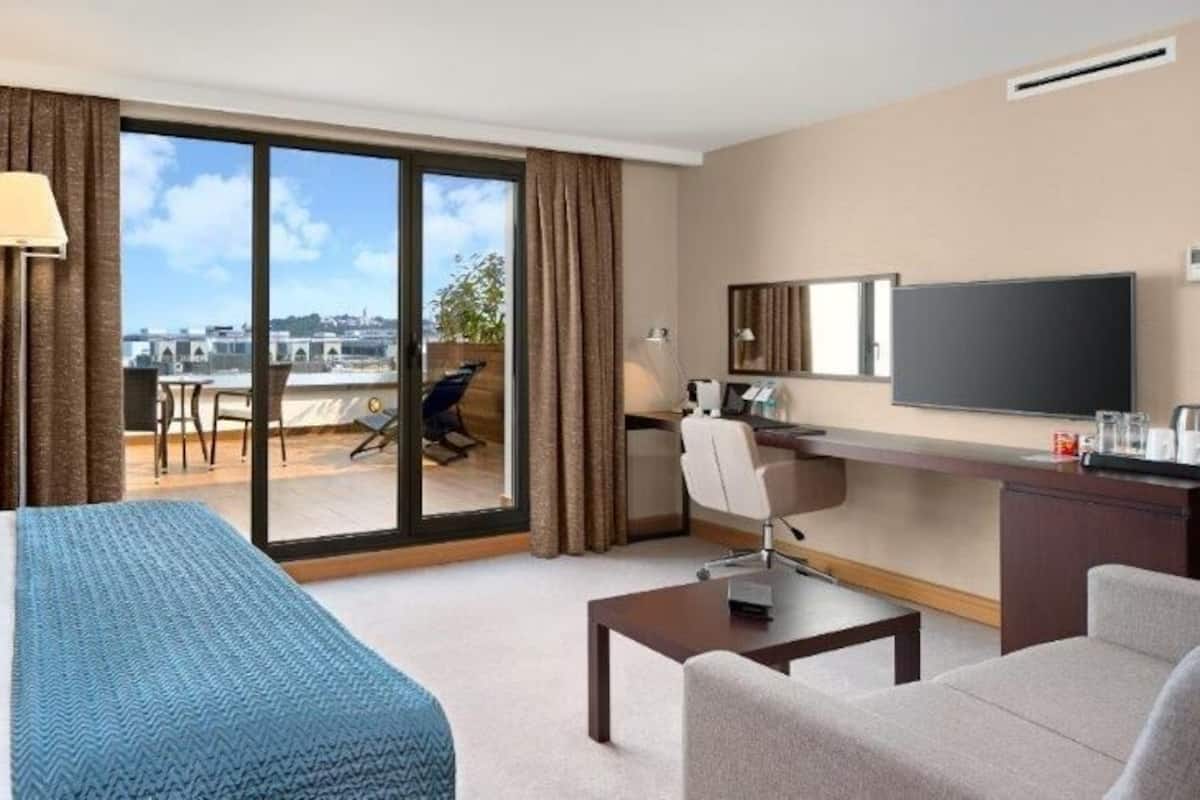 A hotel room with a large bed, a desk, a chair, a television, and a balcony with a view of the city.