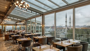 Un restaurante moderno con amplios ventanales que ofrecen vistas a un paisaje urbano y una torre destacada.