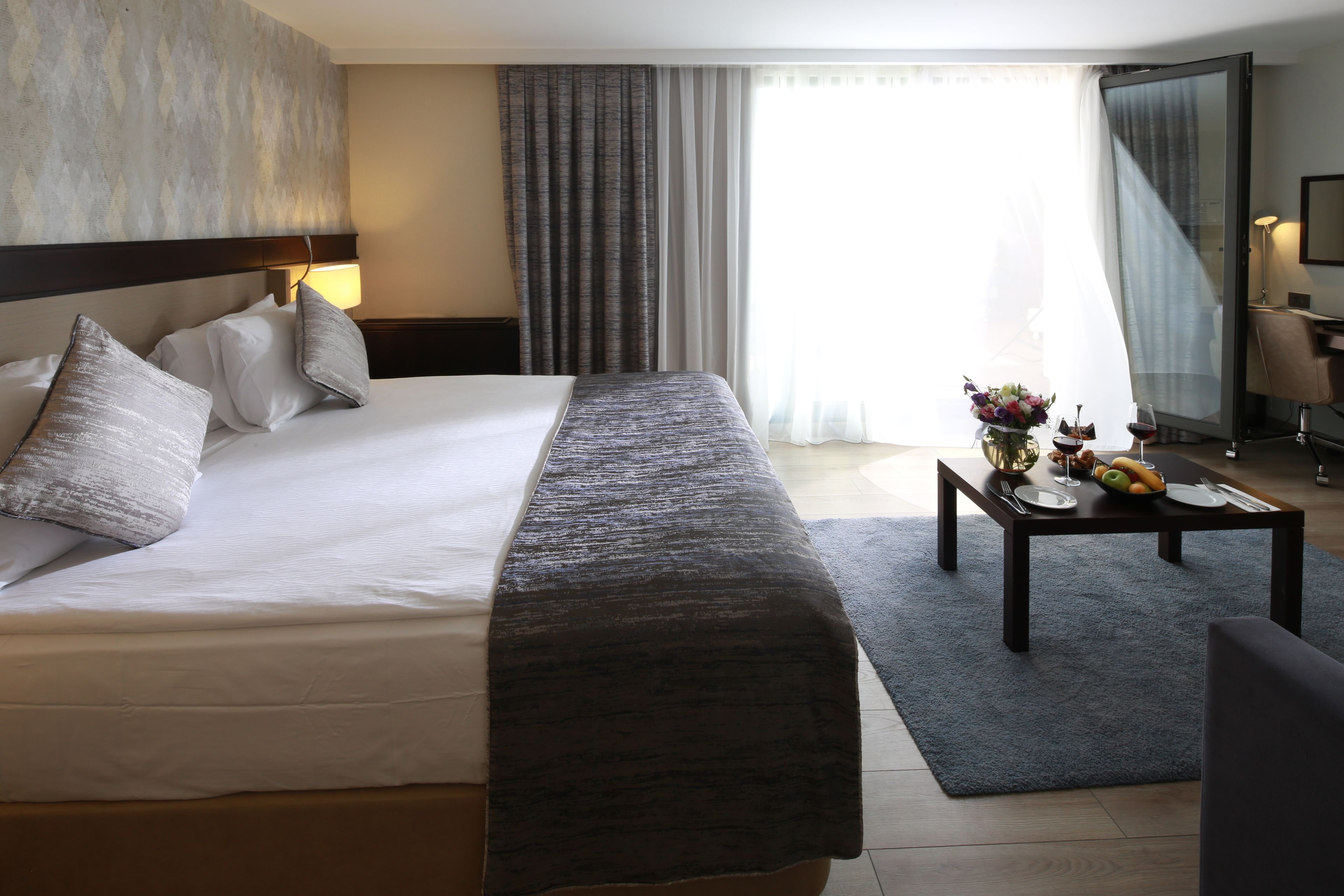 A hotel room with a large bed, a bedside table with a vase of flowers, and a dining table set for two.