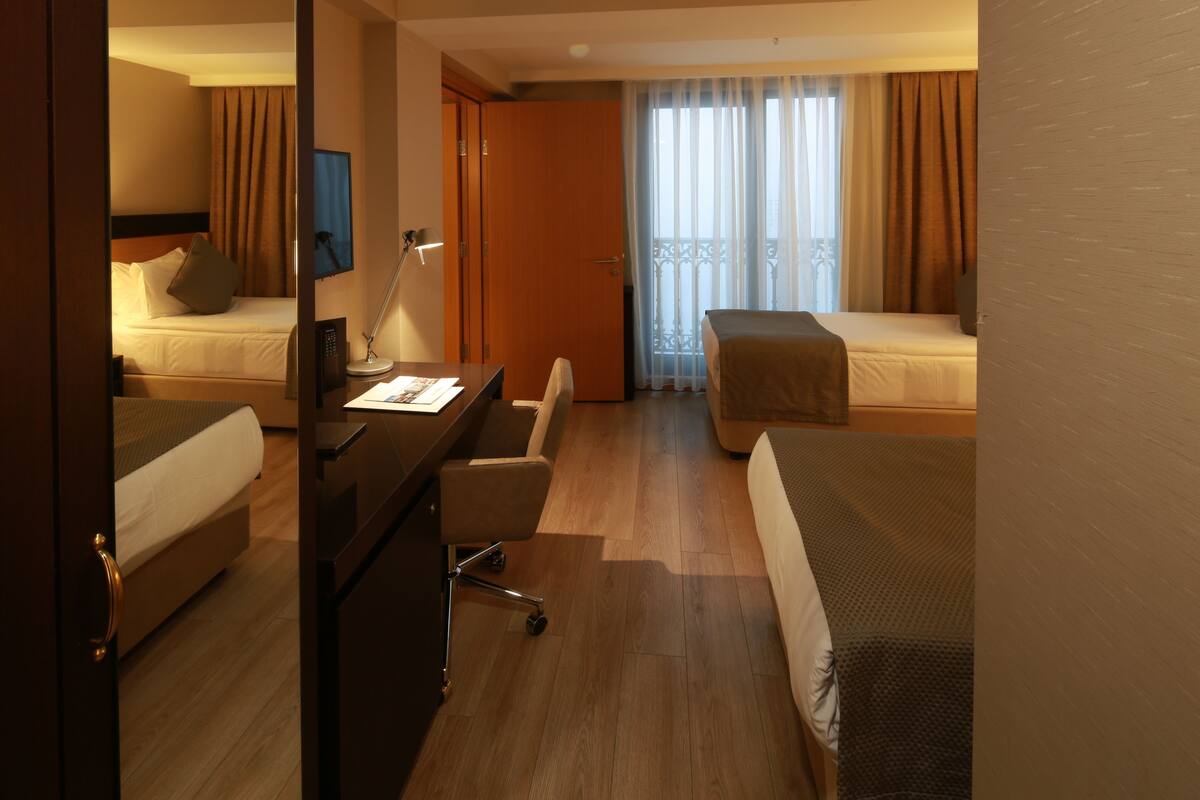 A hotel room with two single beds, a desk with a chair, a TV, and a balcony with curtains.