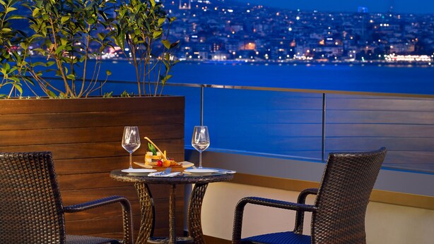 Una mesa de comedor con dos sillas de mimbre y una planta en maceta, con vista a un paisaje urbano nocturno.