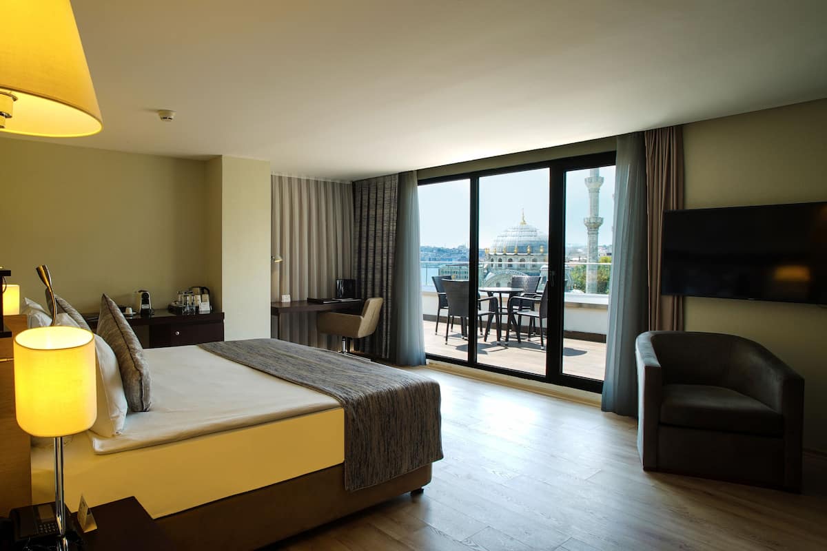 A hotel room with a bed, a desk, a chair, a TV, and a balcony with a view of a cityscape.