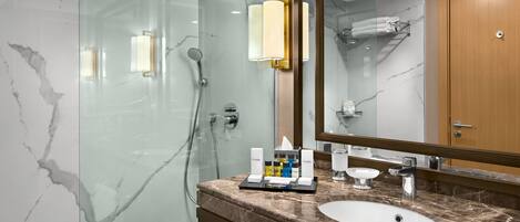 Un baño moderno con ducha envuelta en vidrio, paredes de mármol, un espejo grande y un tocador de mármol con artículos de aseo.
