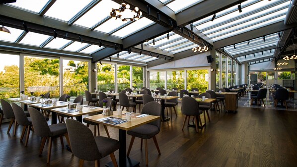 Un restaurant spacieux, doté de hauts plafonds et de grandes fenêtres offrant une vue sur la verdure. Les tables sont disposées avec soin et accompagnées de chaises, et l’intérieur est agrémenté d’un éclairage moderne.