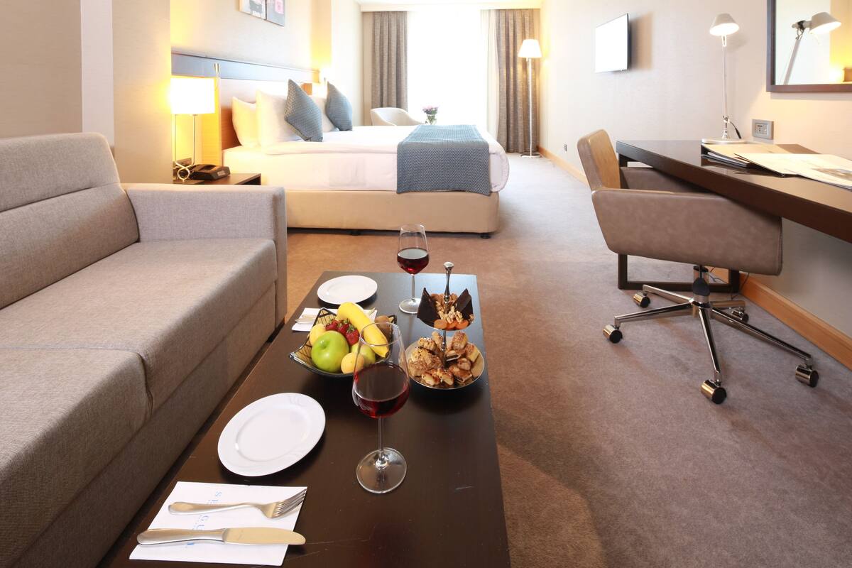 A hotel room with a bed, a sofa, a desk, and a dining table set with plates, cutlery, a glass of red wine, and a fruit bowl.