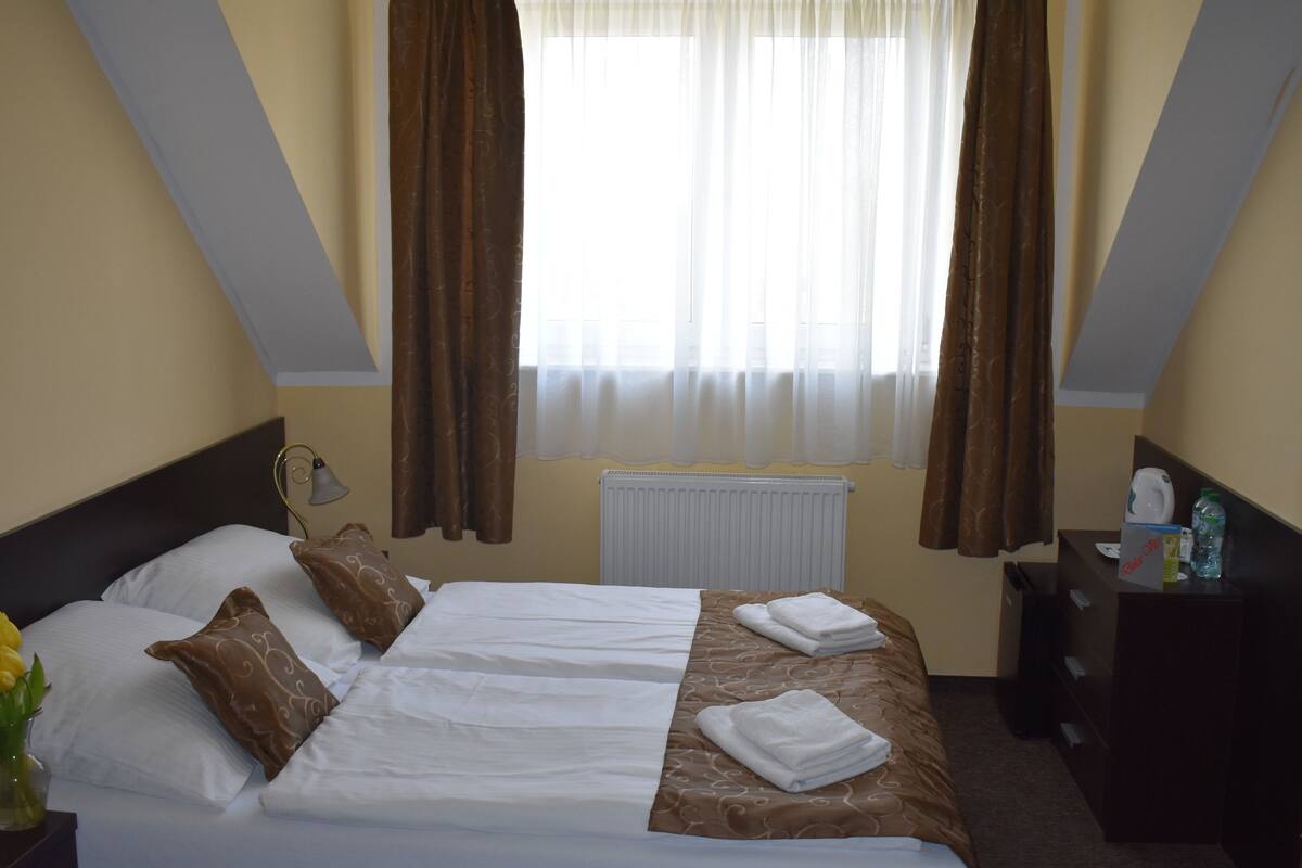 A hotel room with a bed, two pillows, a vase with flowers, a bedside table, a lamp, a radiator, and a window with curtains.