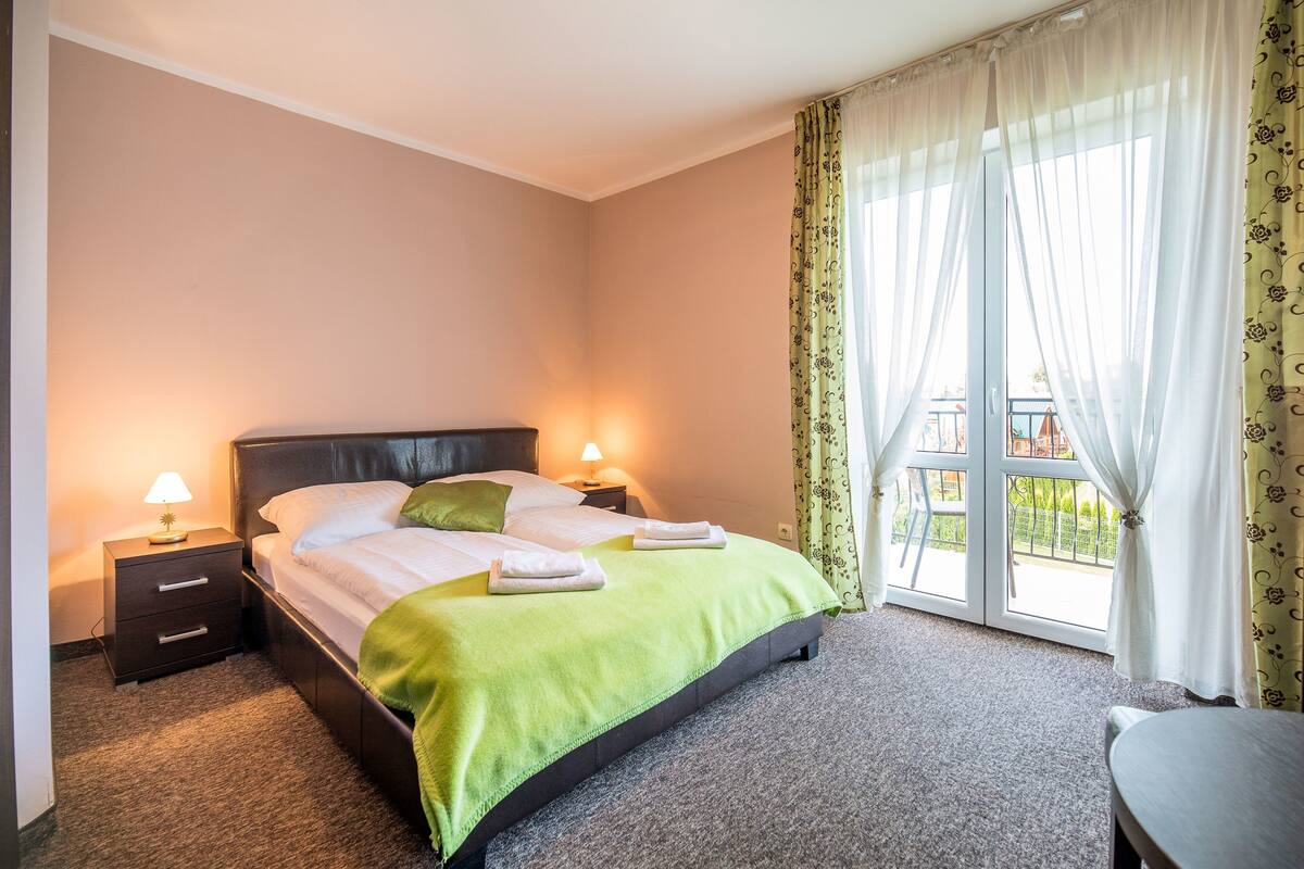 A neatly made bed with a green bedspread, two bedside lamps, and a view of a balcony through sheer curtains.