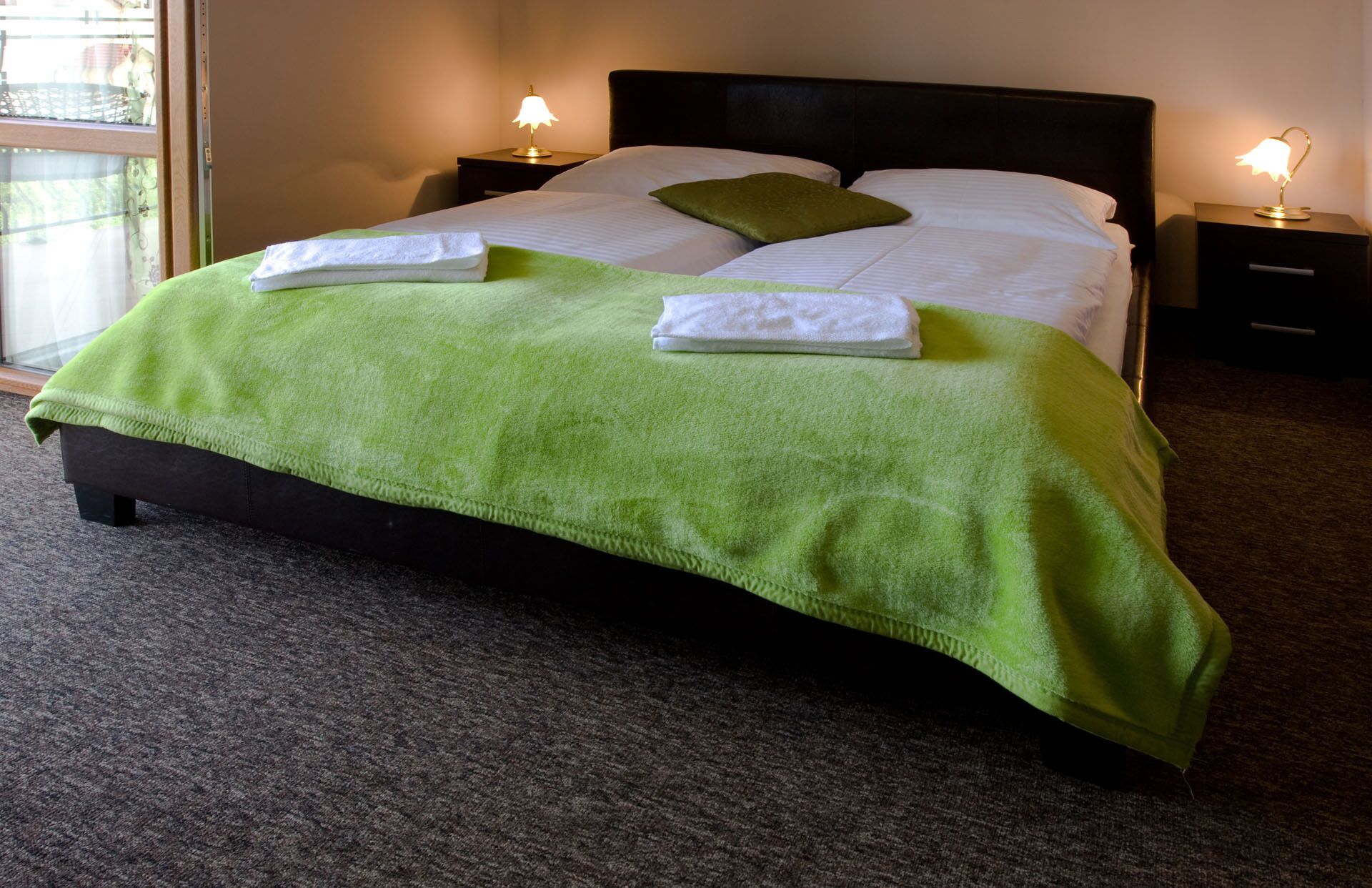 A neatly made bed with a green blanket and white pillows, two bedside lamps, and a view of a balcony through a window.