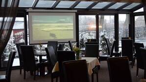 A restaurant with a large projection screen displaying a landscape, black chairs, a table with a white tablecloth, and a view of a snowy outdoor area through large windows. - Dolce  Vita (Ustronie Morskie)