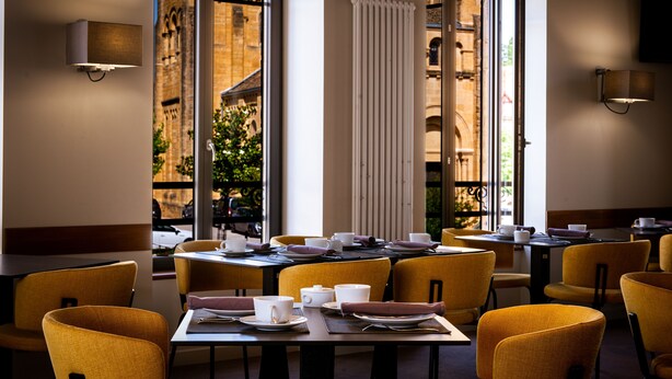 Comedor con mesas dispuestas para el servicio, ventanas amplias con vistas a un edificio histórico y luminarias modernas.