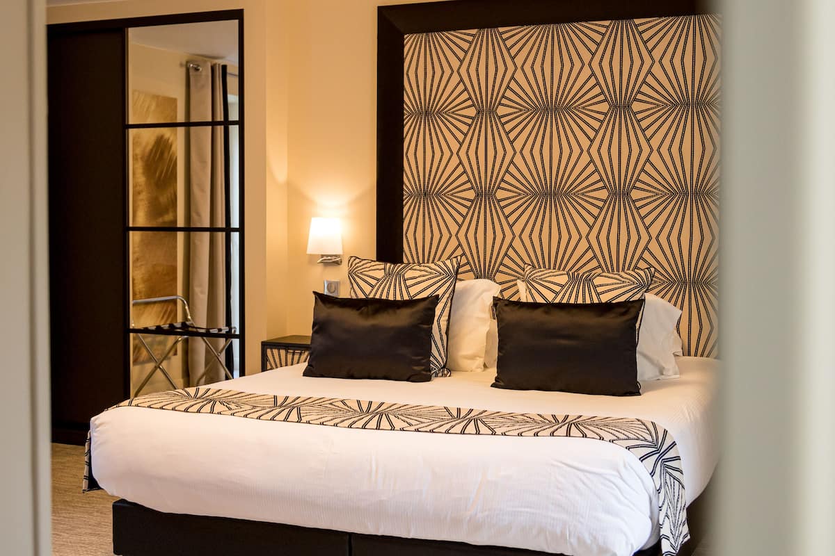 A hotel room with a large bed, a headboard with a geometric pattern, a bedside table with a lamp, and a view of the outside through a door.