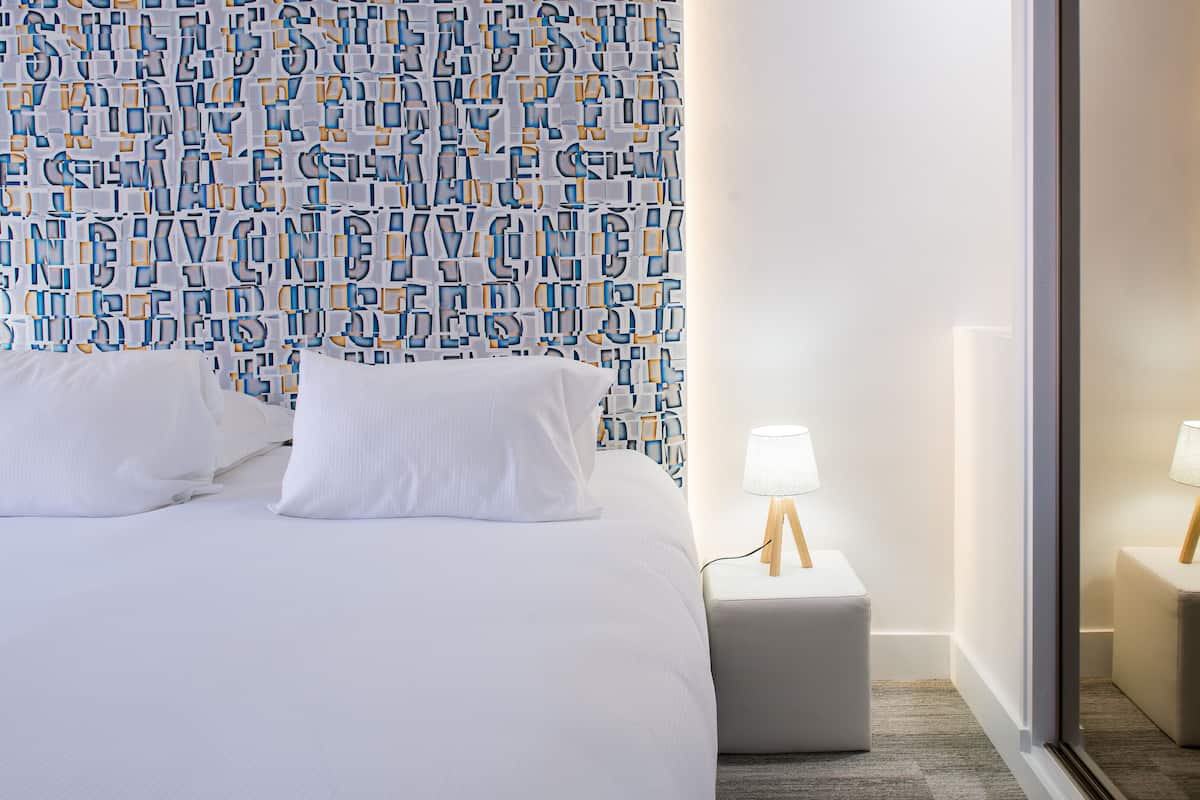 A hotel room with a bed, a nightstand with a lamp, and a mirror reflecting the bed.