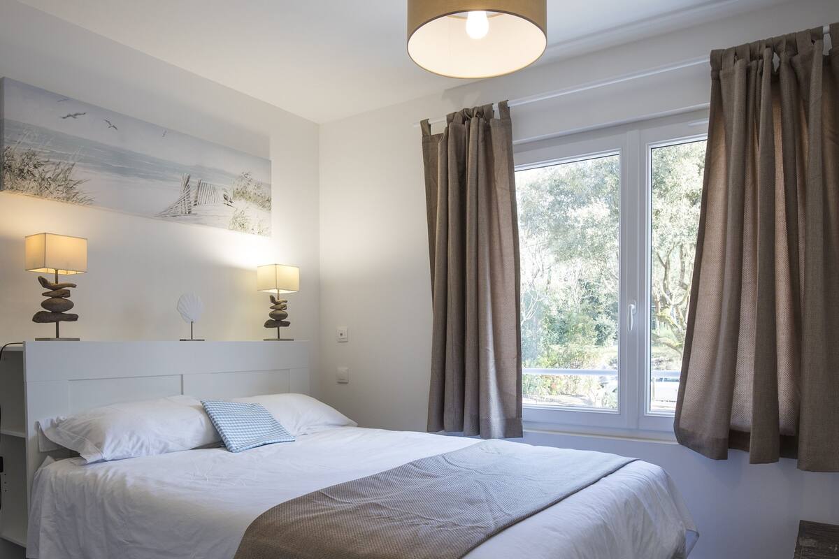Een net opgemaakt bed met witte lakens, een beige plaid en twee bedlampen met lampenkappen.