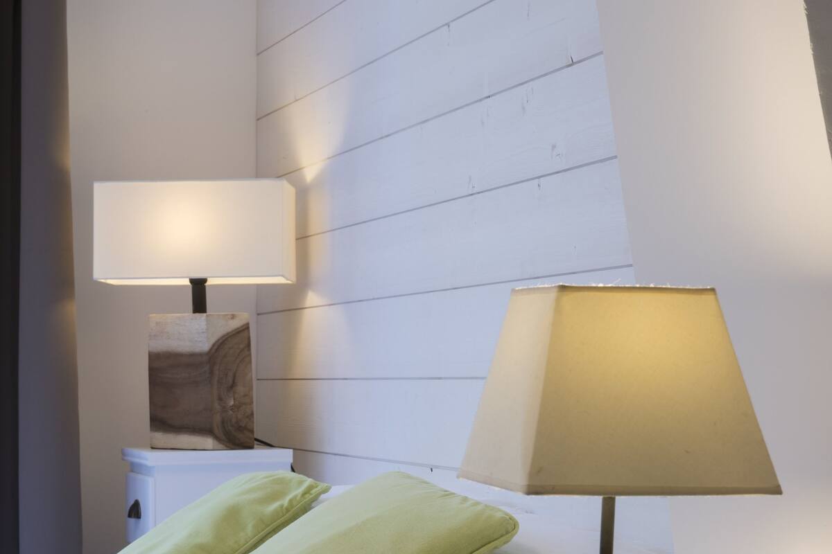Een keurig opgemaakt bed met groene en witte kussens, een nachtkastje met een lamp, en een witte nachtkastje met een fotolijst.