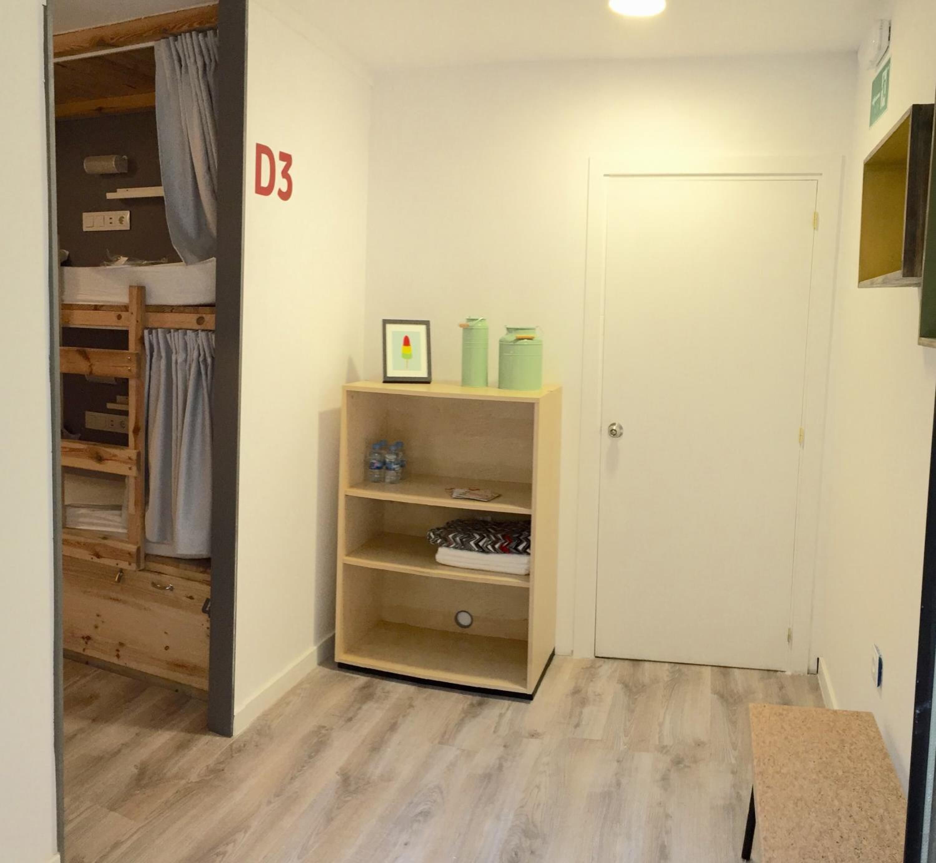 Een gang met een houten stapelbed, een plank met een lijst eromheen en een deur met de aanduiding D3.