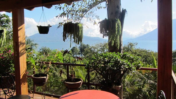 A terrace with potted plants, a table, and chairs, overlooking a mountainous landscape and a body of water. - Casa Hagan (Santa Cruz La Laguna)