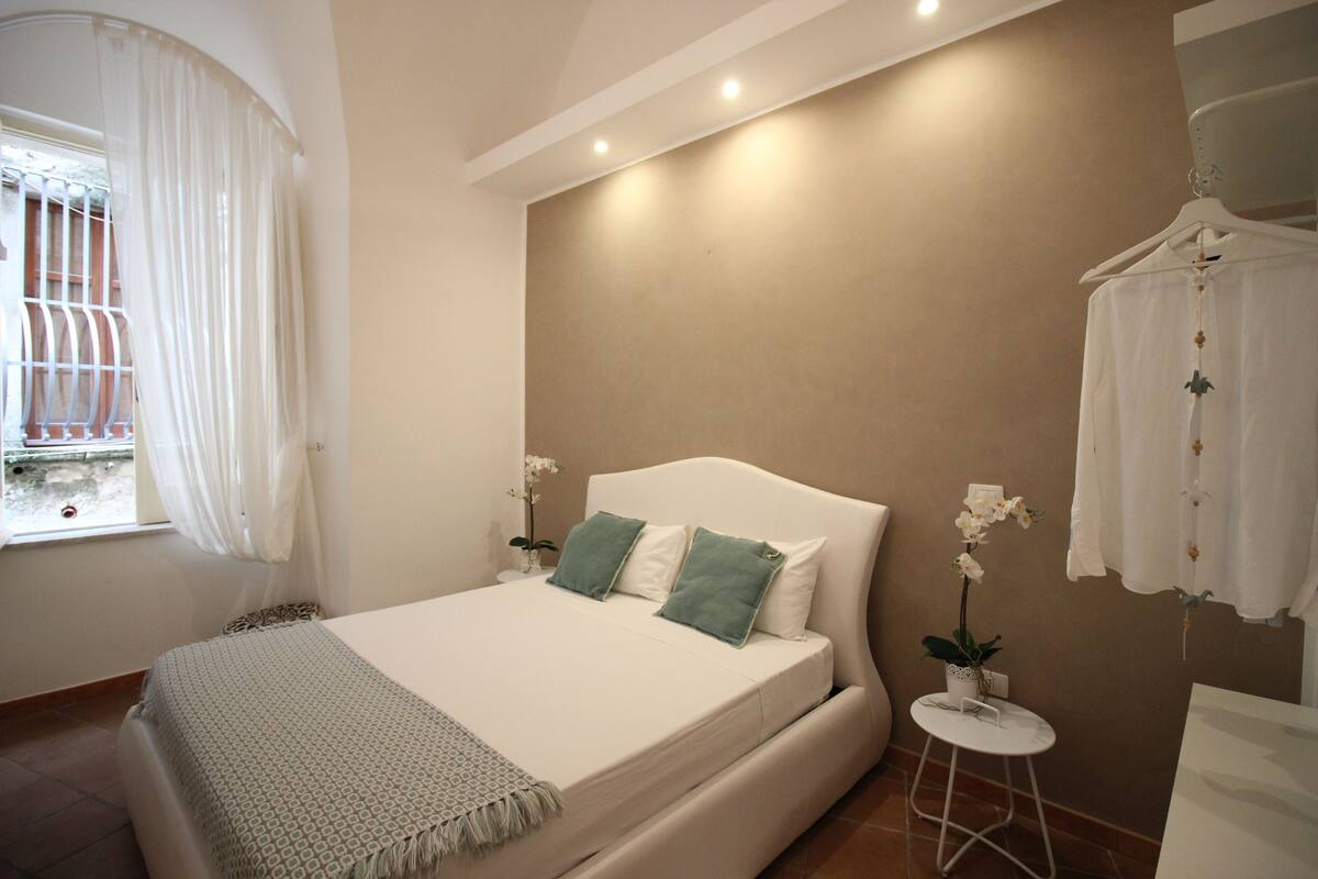 A bedroom with a bed, a bedside table, a hanging light, and a window with curtains.