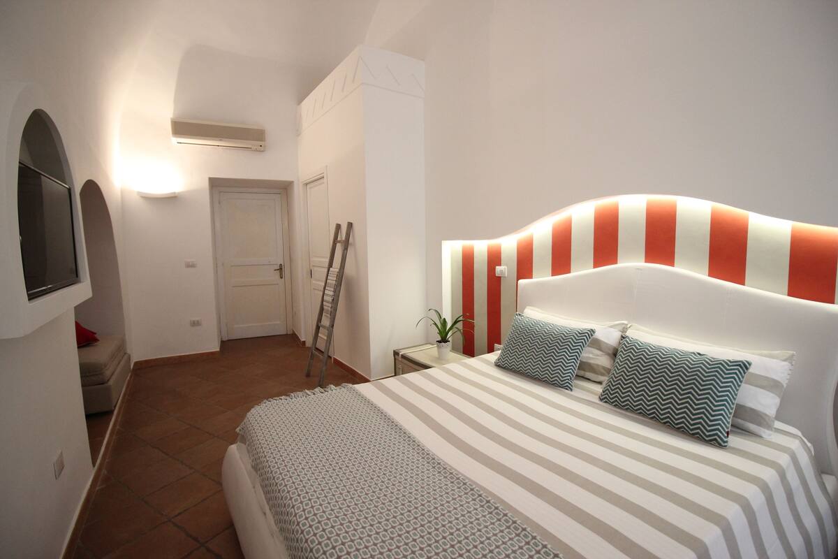 A bedroom with a striped bed, a headboard with orange and white stripes, and a small bedside table with a plant.