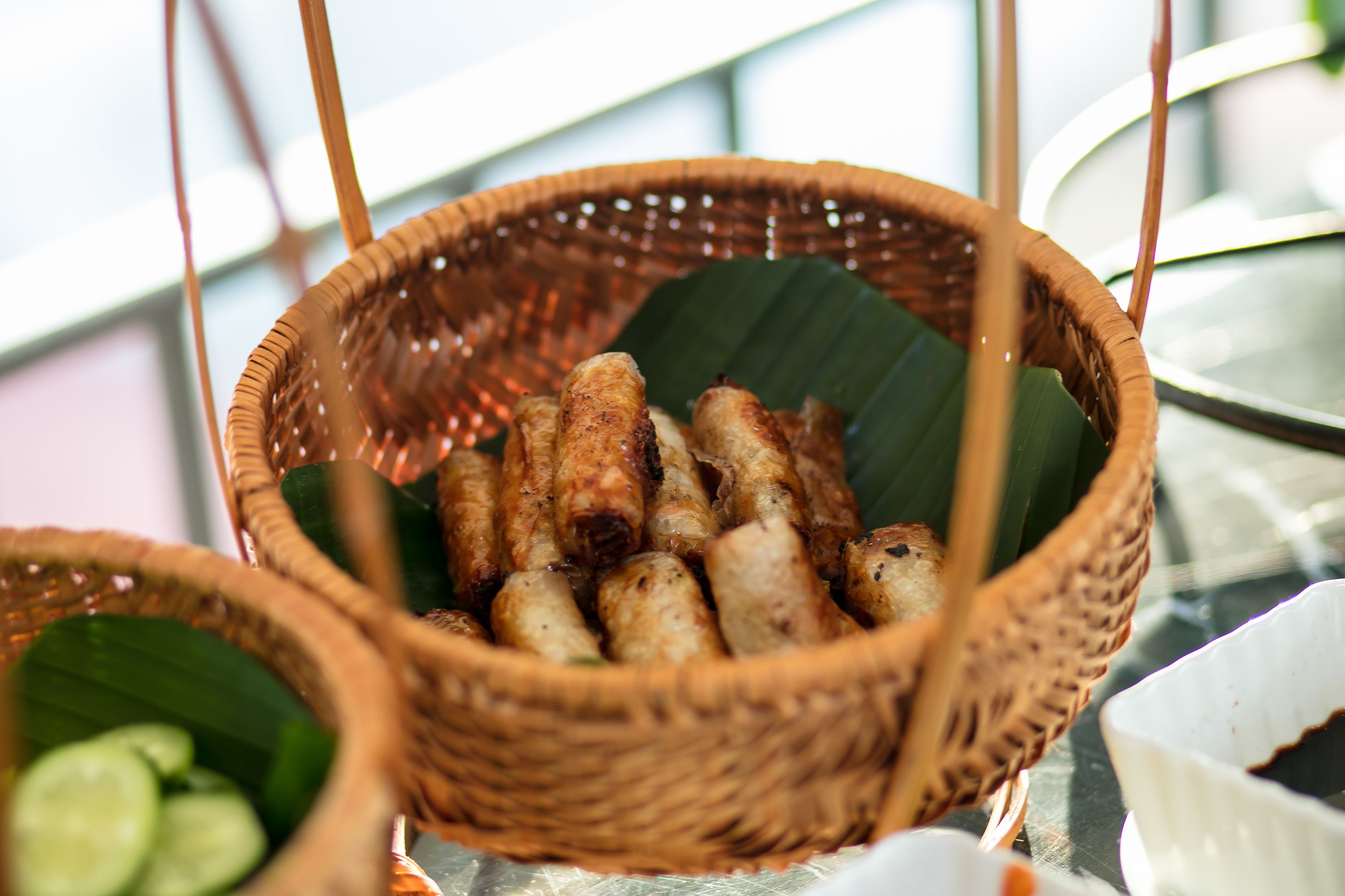 A wicker basket filled with grilled sausages, a bowl of sliced cucumbers, and a white bowl with a dark liquid, possibly soy sauce, on a table.