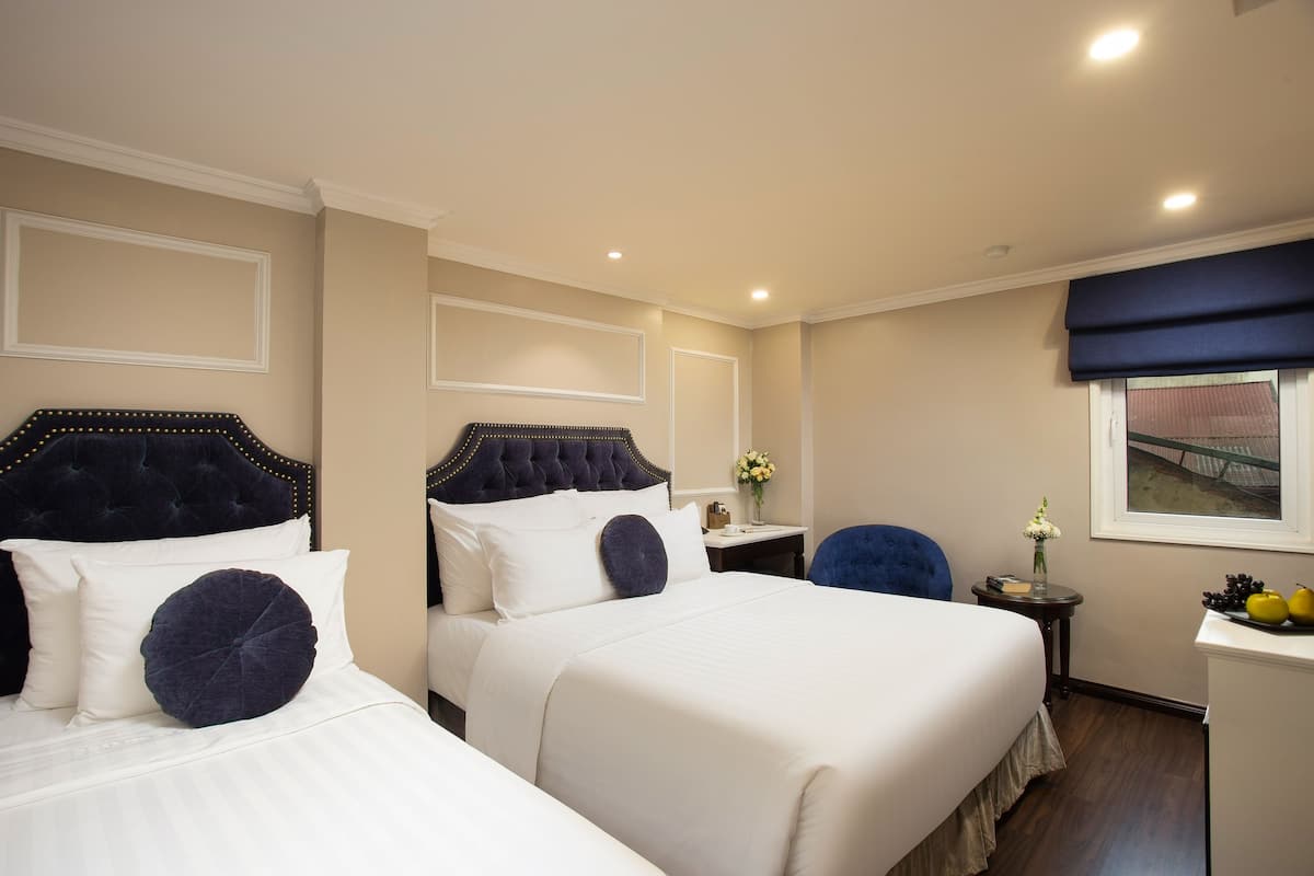 Een hotelkamer met twee bedden, een kleintje tafeltje met een bloemenvaas, een stoel en een raam met uitzicht op een gebouw.
