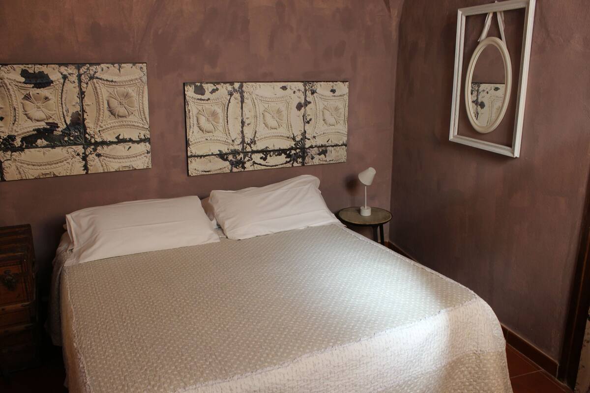 A neatly made bed with white linens in a room with brown walls. There are two wall-mounted artworks and a small table with a lamp.