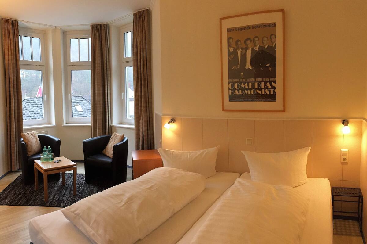 A hotel room with a bed, a chair, a small table with bottles, and a poster on the wall.