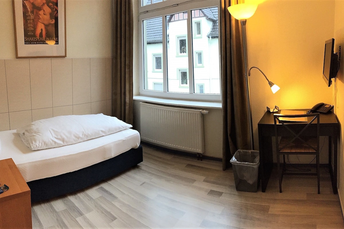 A hotel room with a bed, a desk with a lamp, a trash bin, and a window with a view of buildings.