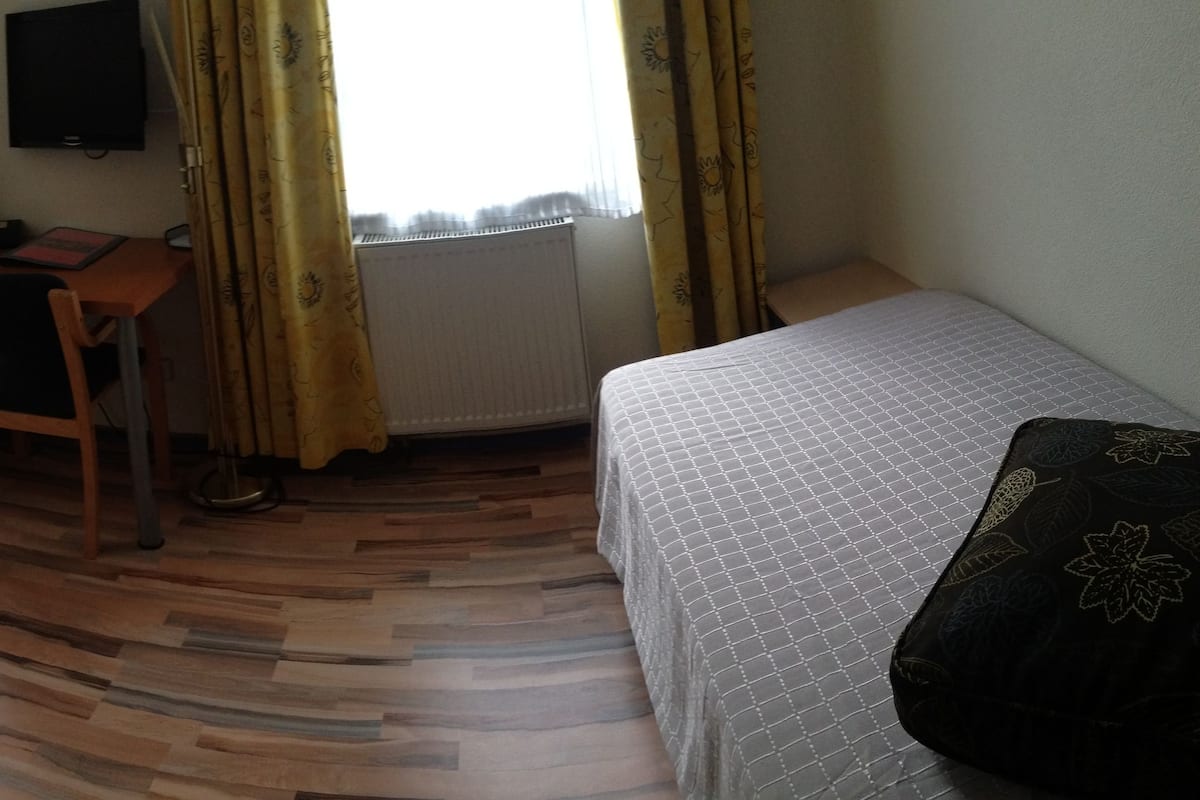 A room with a wooden floor, a bed with a white mattress, a desk with a chair, and a television on the wall.