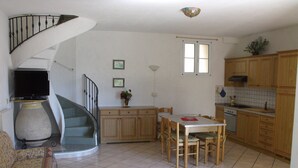 A living room with a sofa, a dining table with chairs, a kitchen area with wooden cabinets, a staircase, and a small window.