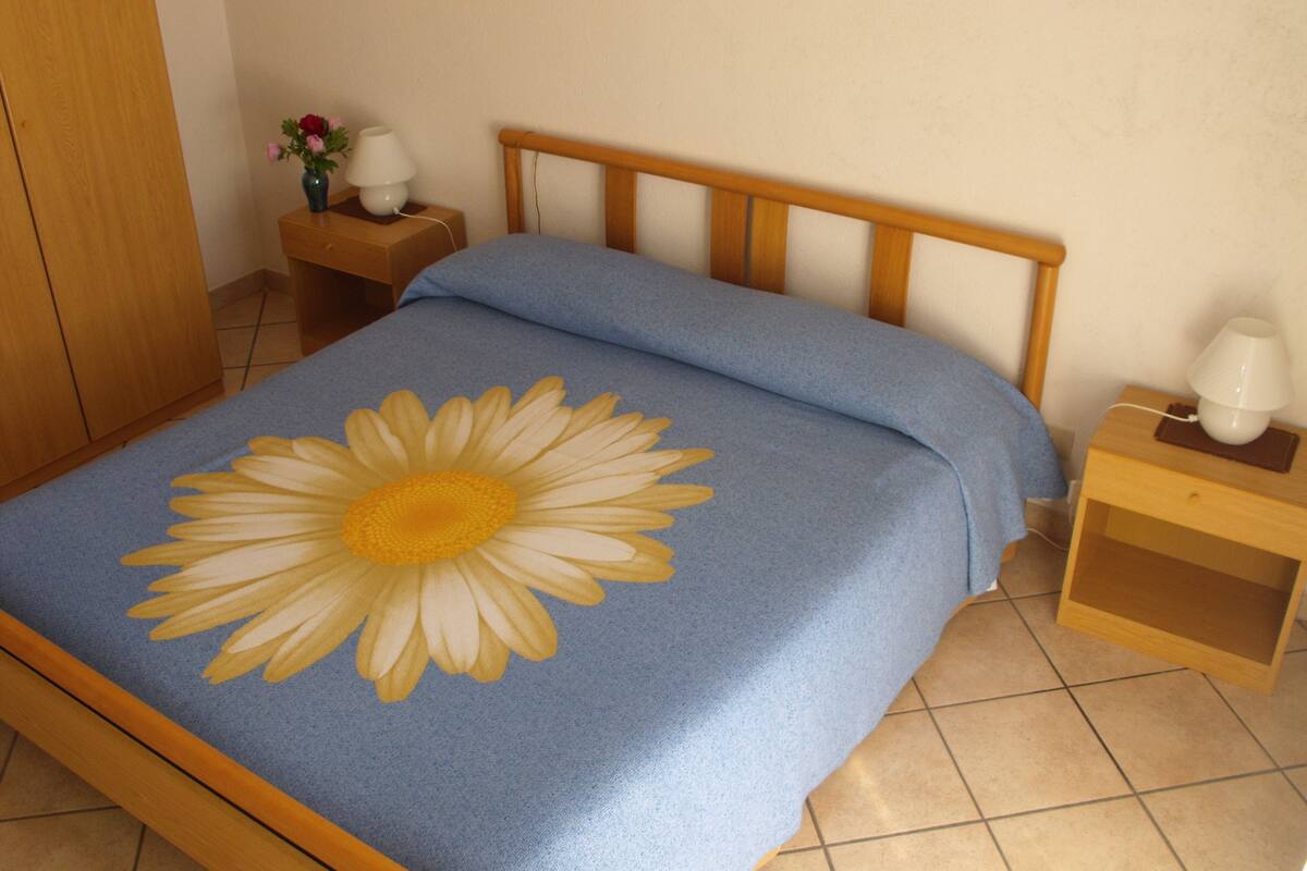 Un dormitorio con cama con colcha estampada de flores, cabecera de madera, mesita de noche con lámpara y armario de madera.
