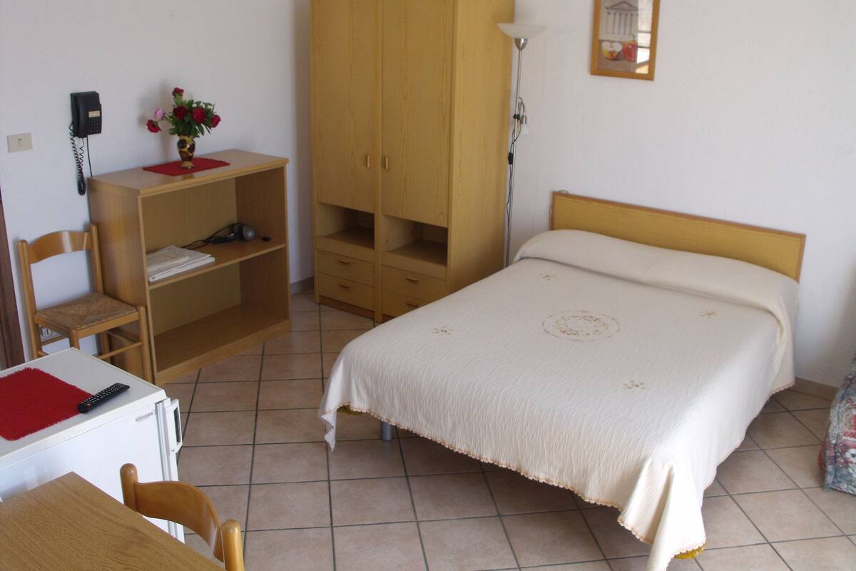 Un dormitorio con cama, armario de madera, escritorio con silla, teléfono y un florero con flores.