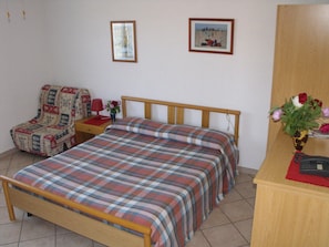 Un dormitori amb un llit de fusta, una cadira, una tauleta de nit, un vas amb flors i dos quadres emmarcats a la paret.