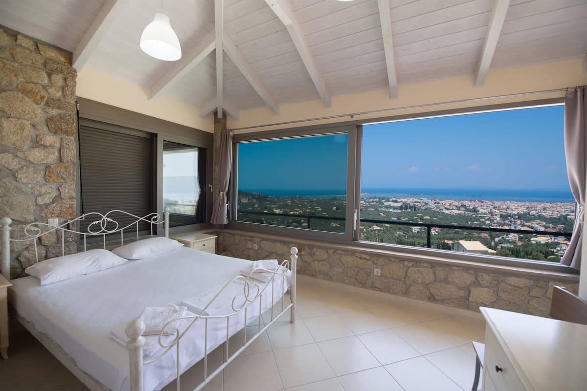 A bedroom with a large window, a bed, a nightstand, and a stone wall.