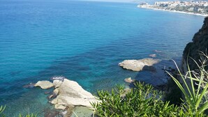 A coastal landscape with clear blue waters, rocky outcrops, and a distant town. - B&B Il Nido (Ricadi)