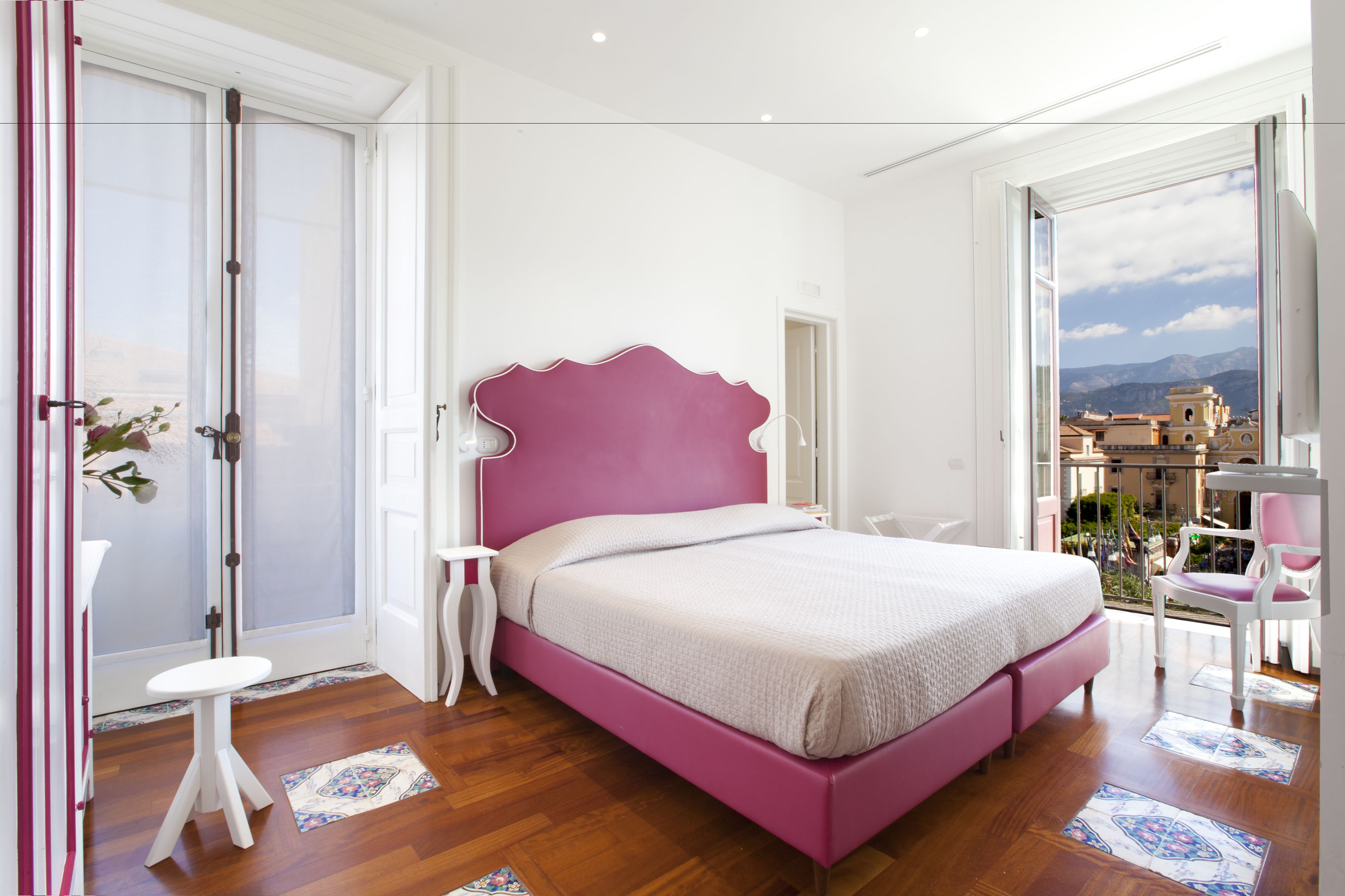 A bedroom with a pink bed, white walls, and a balcony with a view of buildings and mountains.