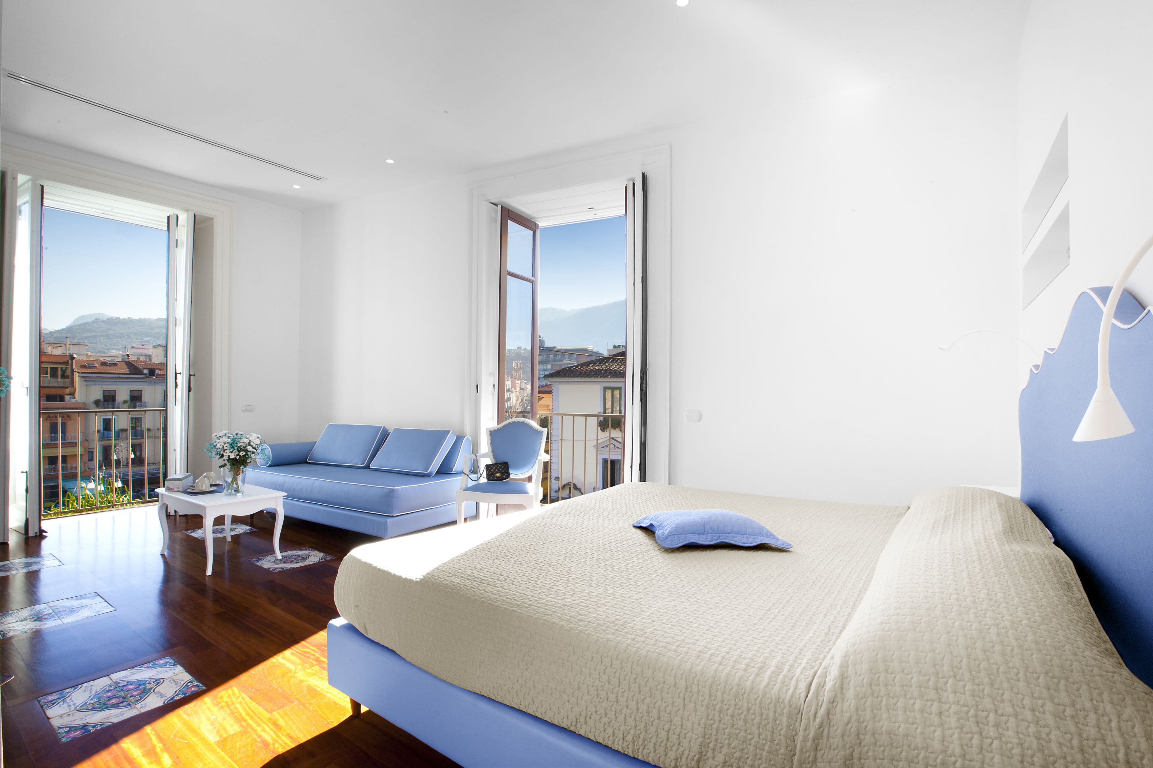 A modern hotel room with a large bed, a blue sofa, a small table, and a balcony with a view of the city.