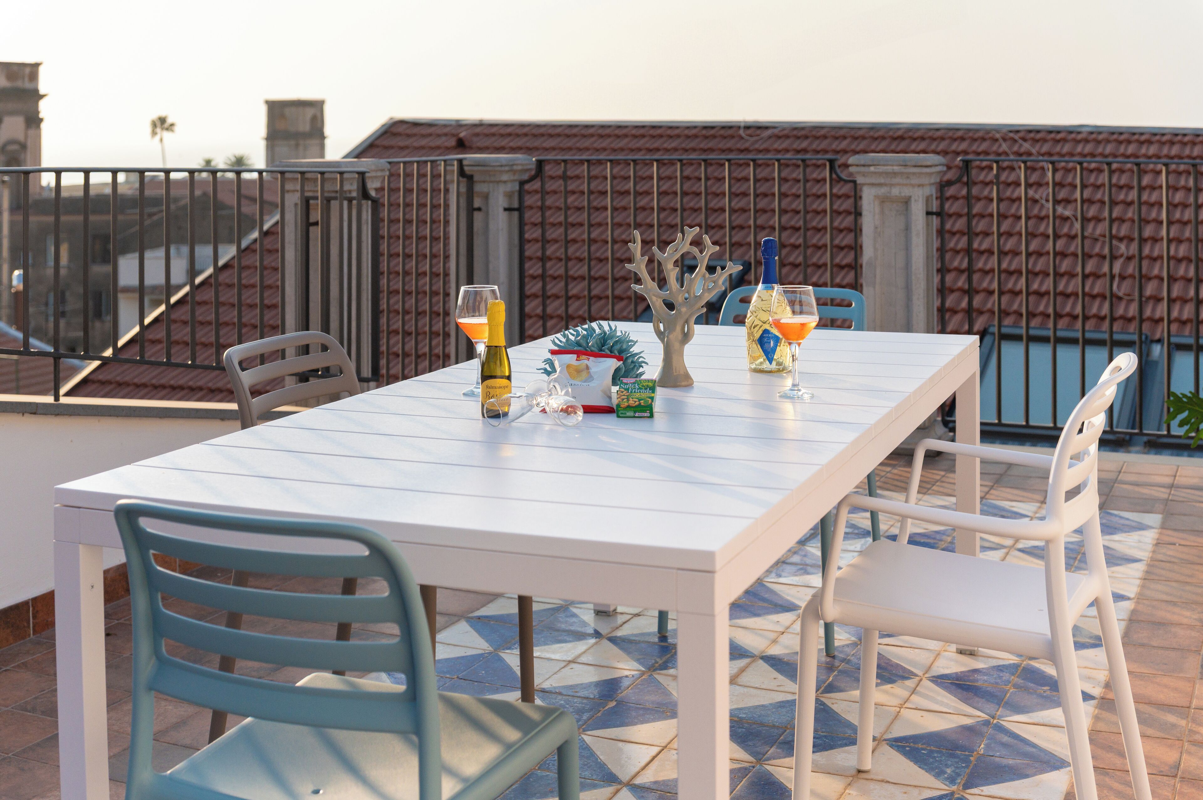 Une terrasse avec une table blanche dressée pour deux personnes, agrémentée d’une bouteille de vin, d’un ananas et d’un objet décoratif en forme de corail.