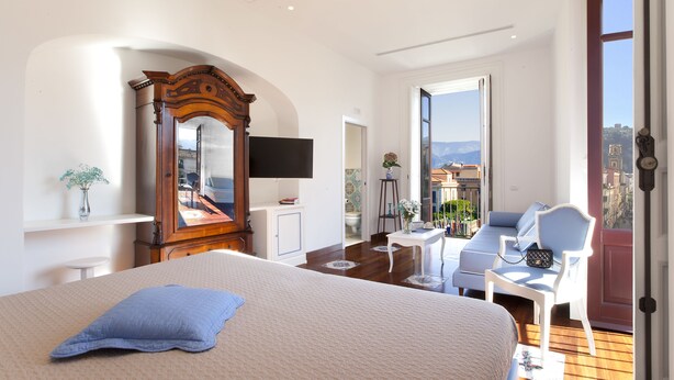 Une chambre à coucher comprenant un lit, une commode, un miroir, une télévision, un balcon avec vue sur les immeubles et une fenêtre.