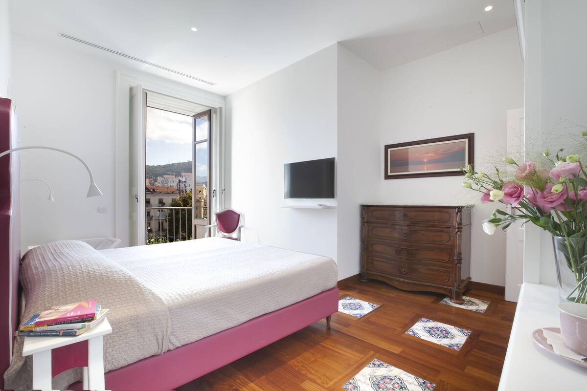 Une chambre à coucher comprenant un lit, une commode, un téléviseur fixé au mur, une chaise et une fenêtre donnant sur des immeubles.