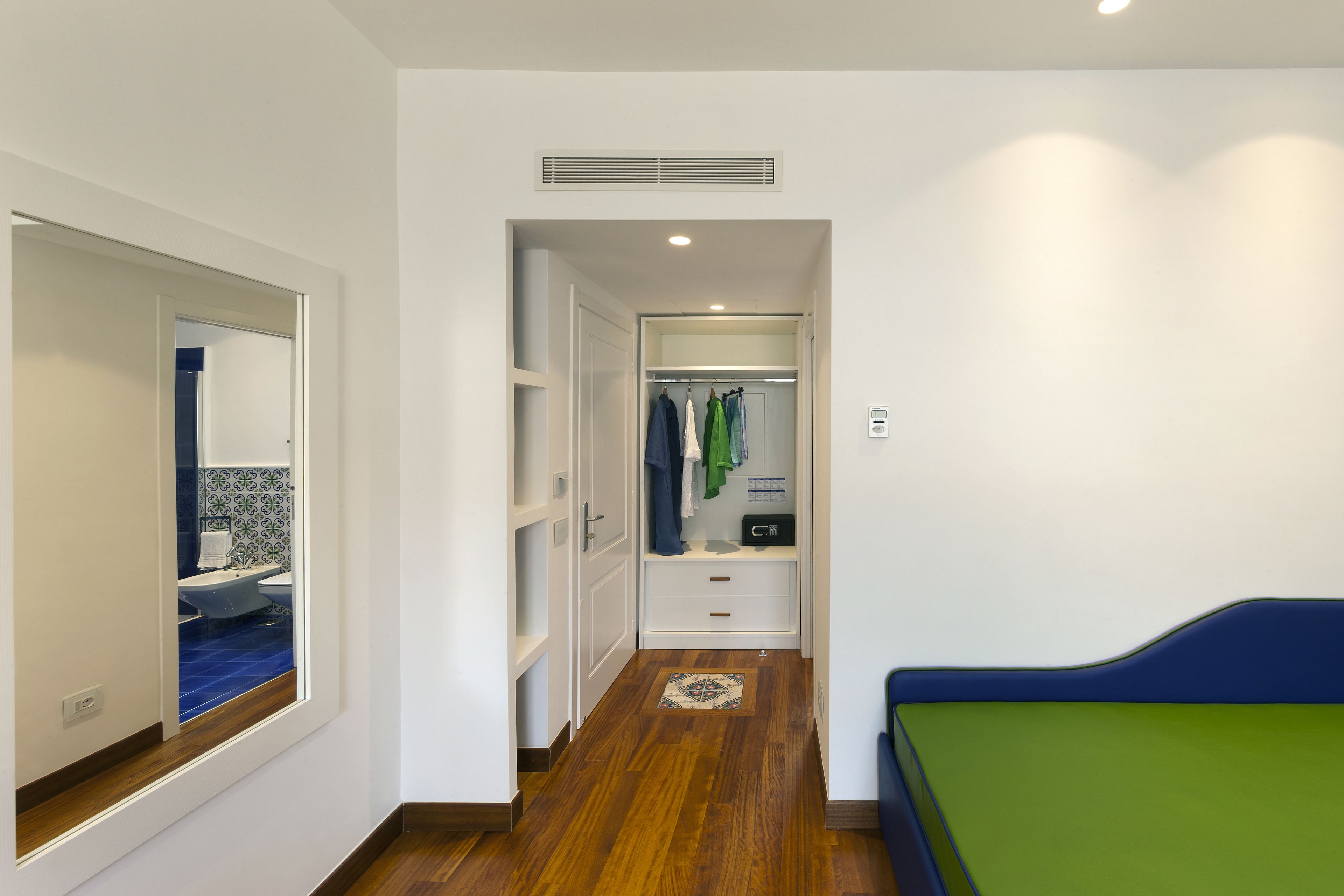 A narrow hallway with a wooden floor, a mirrored closet door, and a bed with a green and blue bedspread.