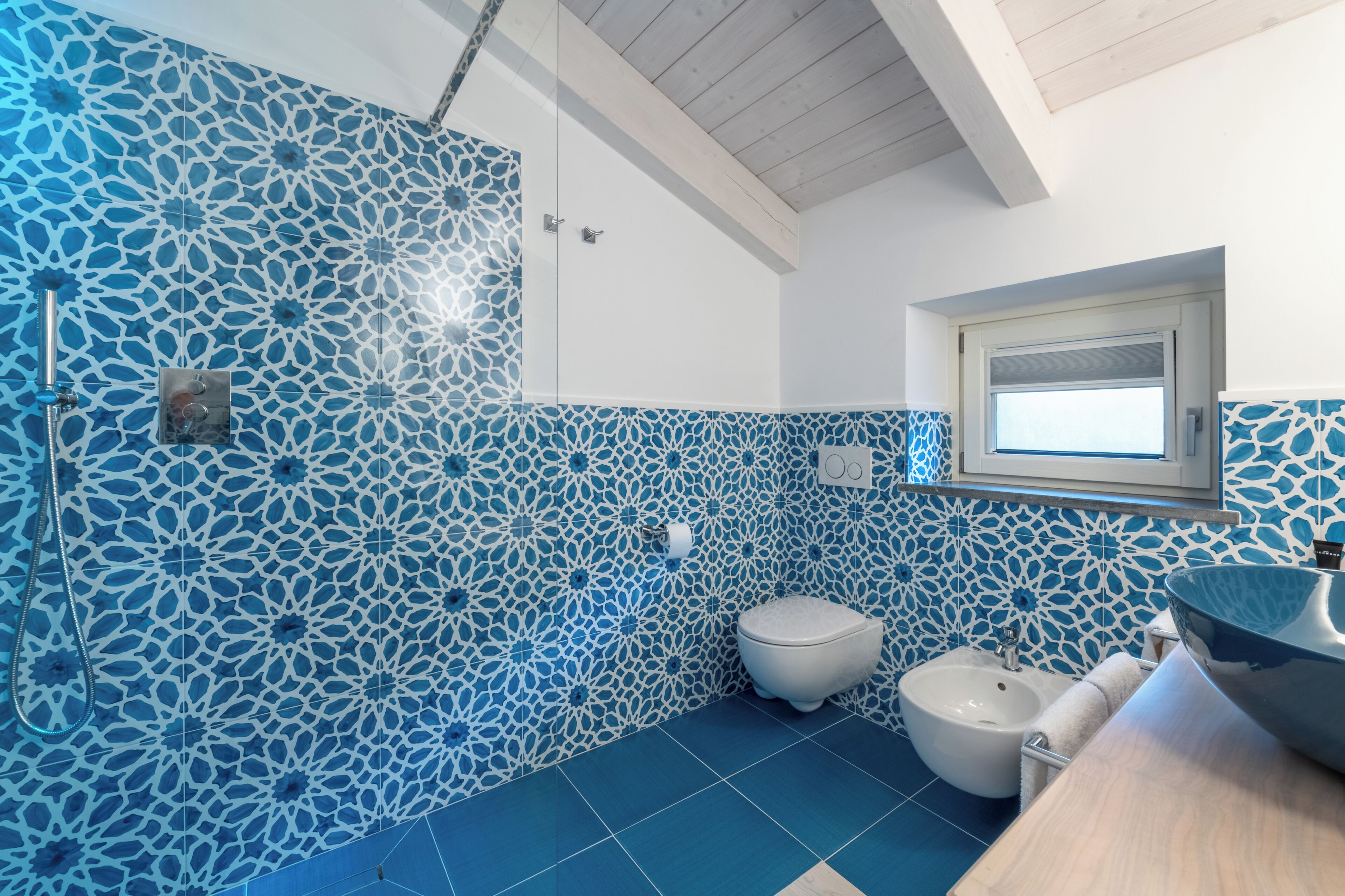 A bathroom with blue patterned tiles, a white toilet, a bidet, and a wooden vanity with a mirror.
