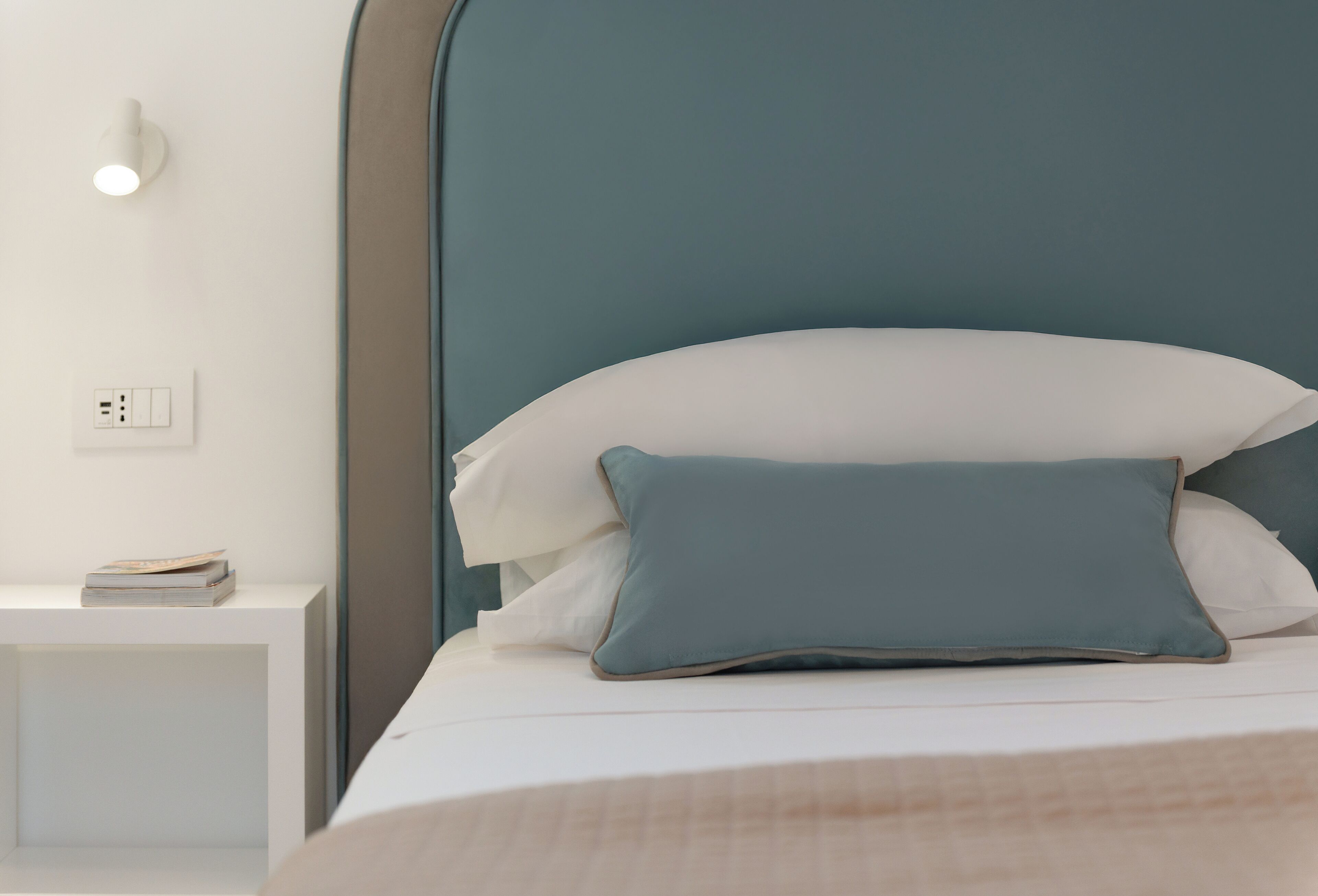 A neatly made bed with a white headboard and two pillows, a bedside table with a book, and a wall-mounted light.