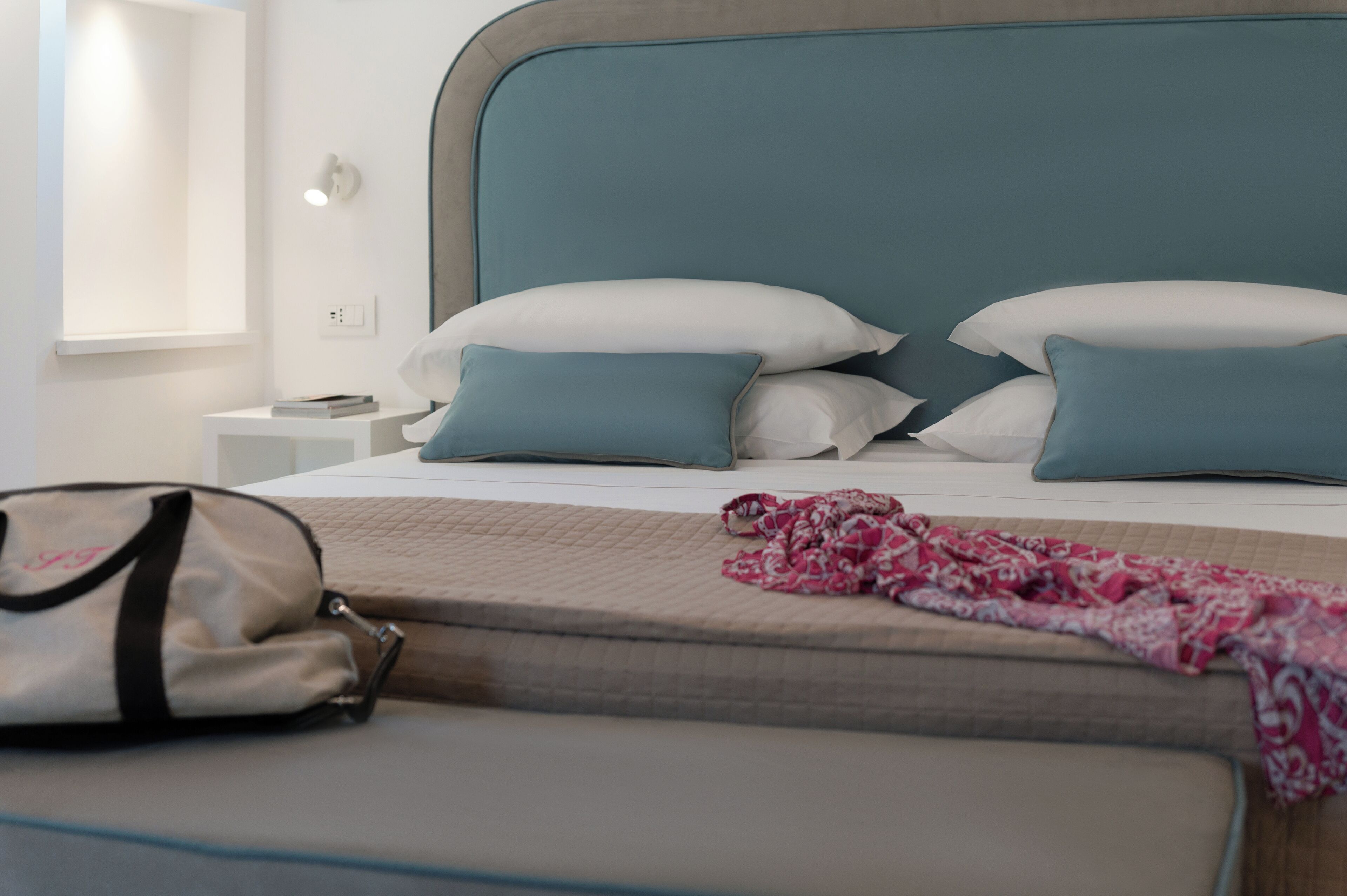 A hotel room with a bed, pillows, a bedside table, a lamp, and a bag on the floor.