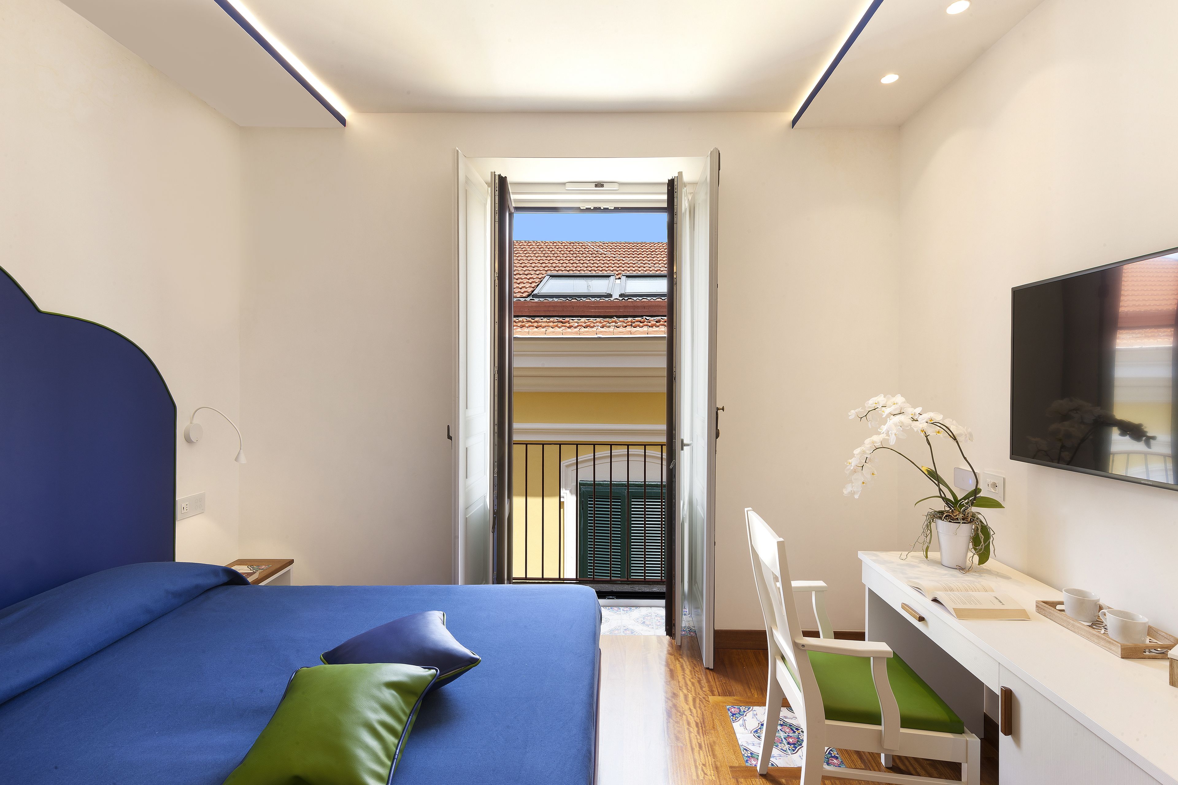 A bedroom with a bed, a desk, a chair, a TV, and a balcony with a view of a building.