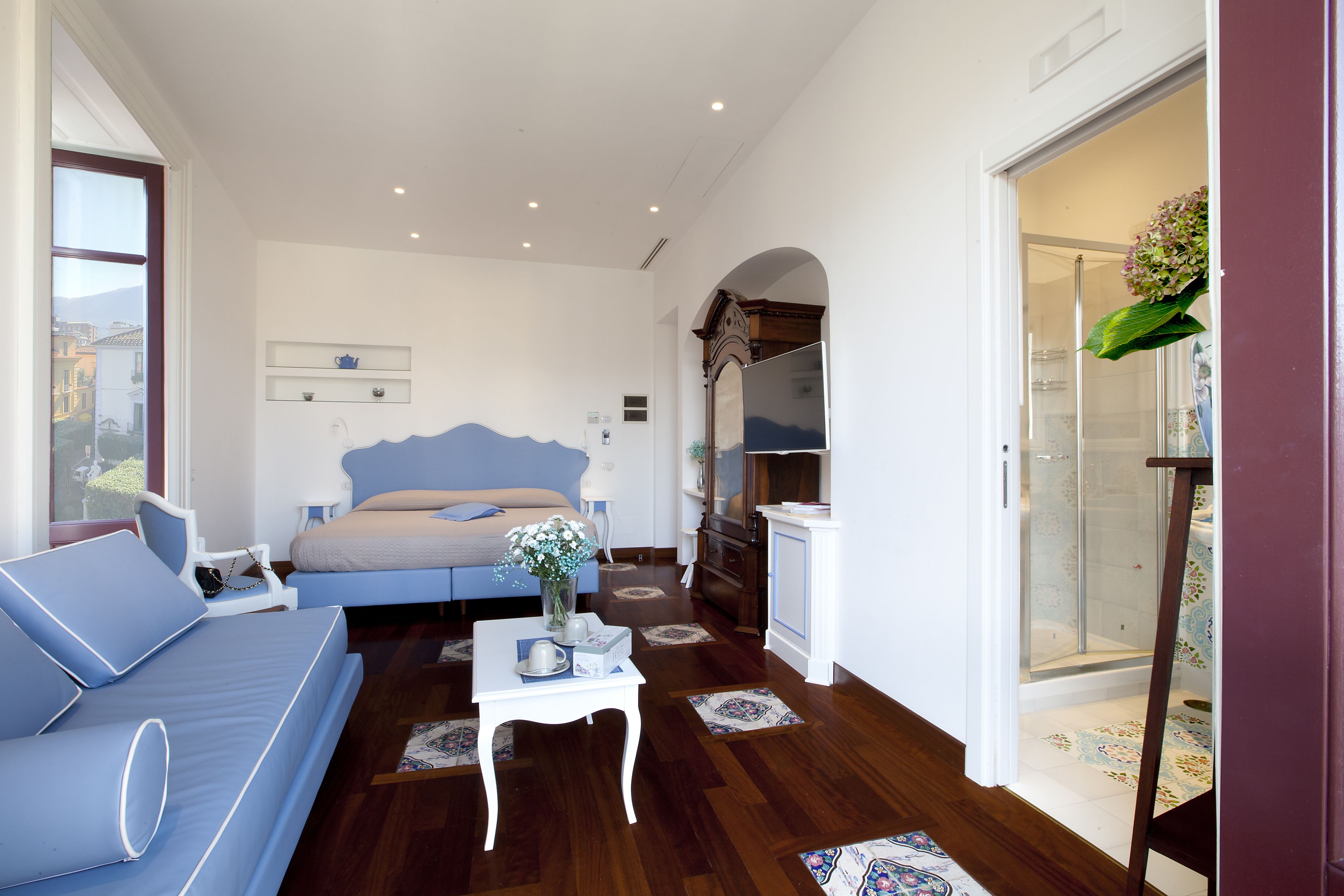A bedroom with a bed, a blue sofa, a small table with a vase, and a shower area.