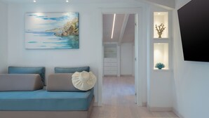 Un salon moderne avec un canapé bleu, un tableau représentant un paysage côtier et un couloir menant à une chambre à coucher.
