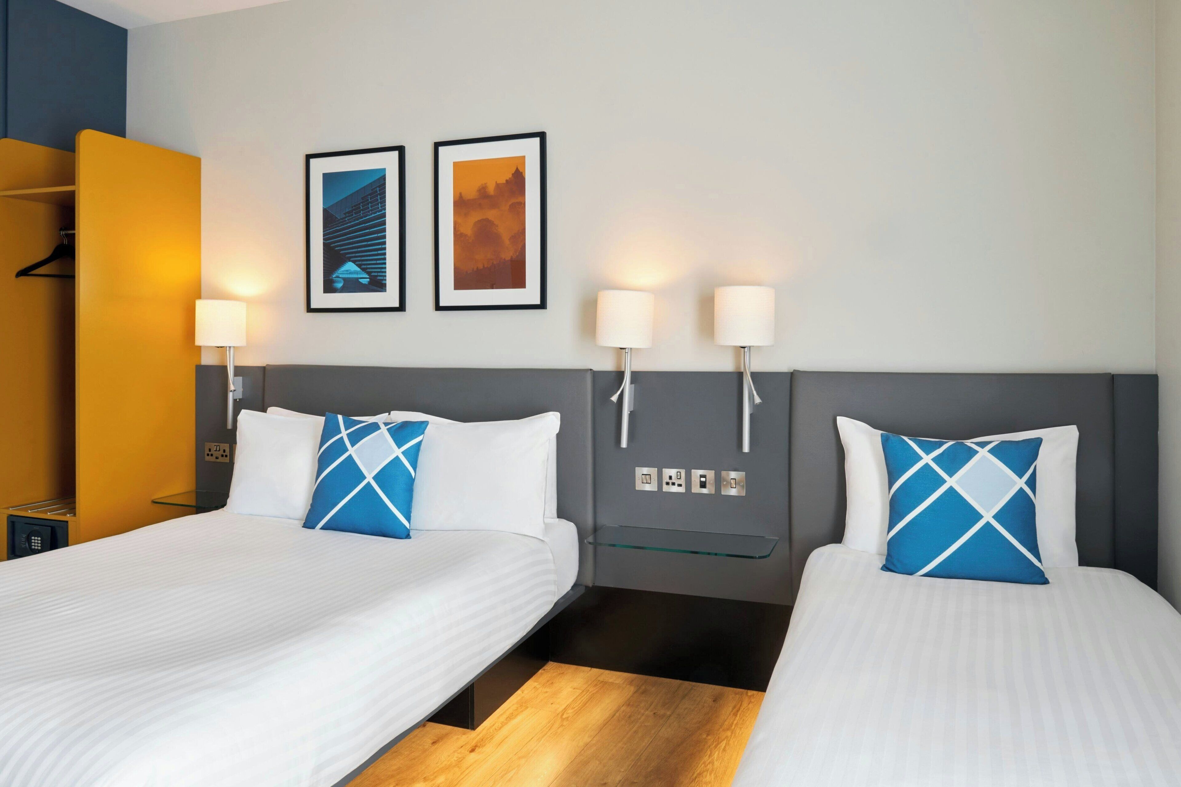A hotel room with two beds, a wardrobe, a bedside table, wall-mounted lamps, and two framed pictures.